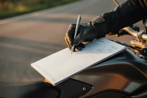 Main gantée d'un motard signant un document d'assurance moto sur le réservoir, en bord de route belge au coucher du soleil.