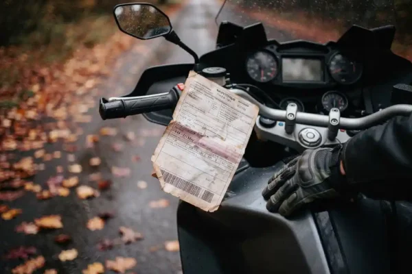 Document d'assurance moto glissé sous le guidon d'un touring garé au bord d'une route belge, feuilles d'automne sur l'asphalte humide.