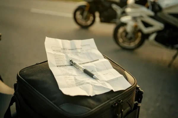 Formulaire de constat amiable froissé posé sur un sac de réservoir moto, stylo bille en diagonale, lumière dorée de fin d'après-midi.