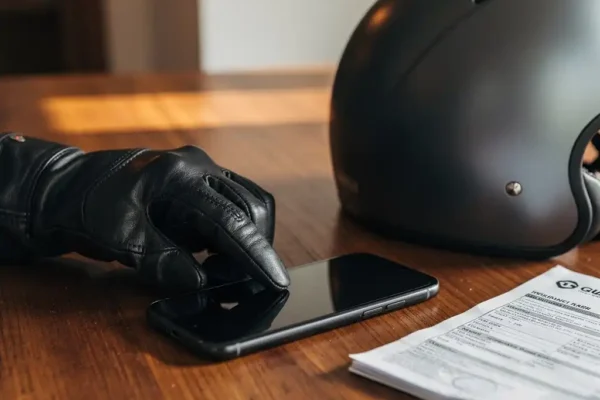 Main gantée de motard mettant à jour ses informations sur smartphone, casque moderne et document d'assurance posés sur un bureau en bois.