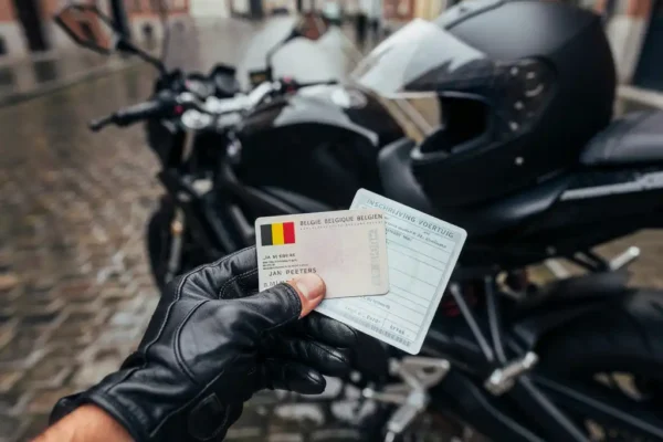 Main portant une carte d'identité belge et un certificat d'immatriculation près d'une moto noire garée sur des pavés.