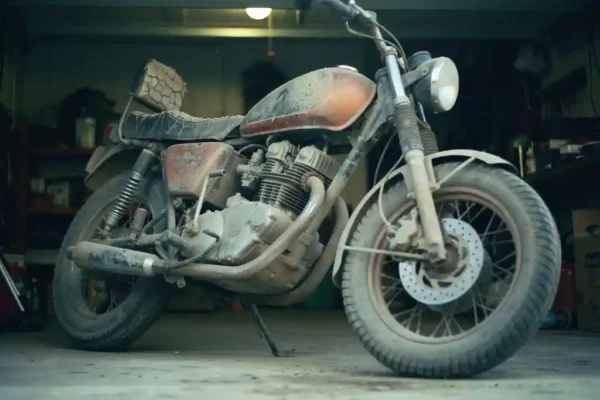 Moto à béquille latérale dans un garage sombre, réservoir poussiéreux, pneus à plat et selle en cuir craquelée témoignant d'un long abandon.