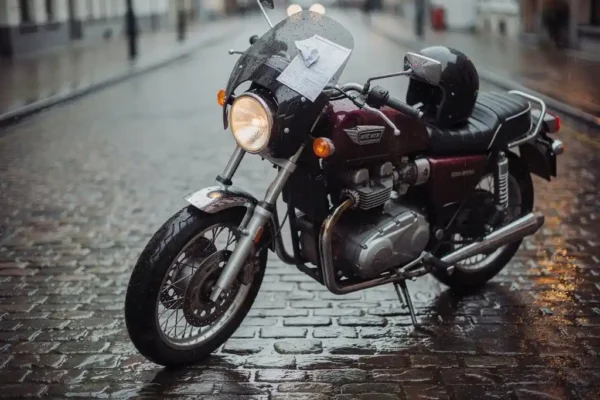 Moto garée sur des pavés belges mouillés, document d'assurance glissé sous le pare-brise, casque posé sur la selle, ciel couvert.