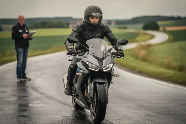 Motocycliste en tenue complète pratiquant des manœuvres lentes sur une route de campagne belge mouillée, sous l'œil d'un instructeur.