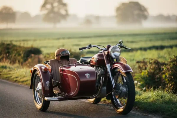 Moto vintage avec side-car bordeaux garé sur une route de campagne belge, brume matinale dorée sur les champs verts, casque en cuir posé sur la selle.