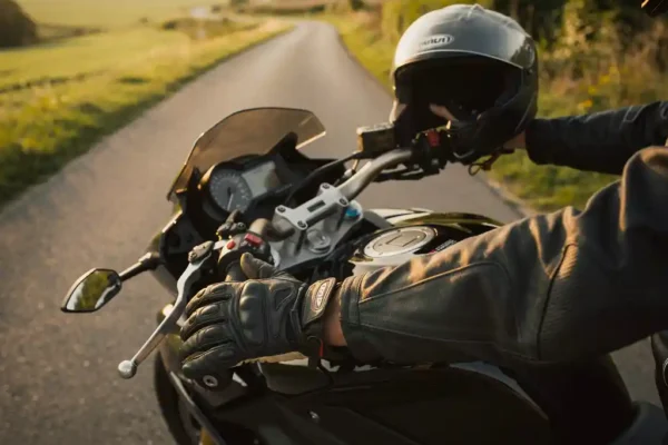 Mains gantées de cuir sur le guidon d'une moto de tourisme garée, casque et veste posés sur la selle, campagne belge en arrière-plan.