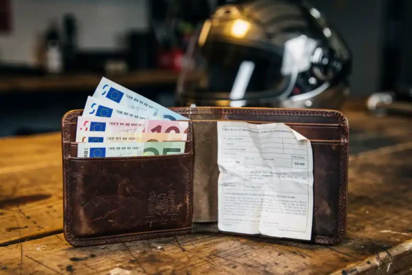 Portefeuille moto en cuir vieilli ouvert sur un établi, contenant des billets en euros et un document d'assurance moto plié.