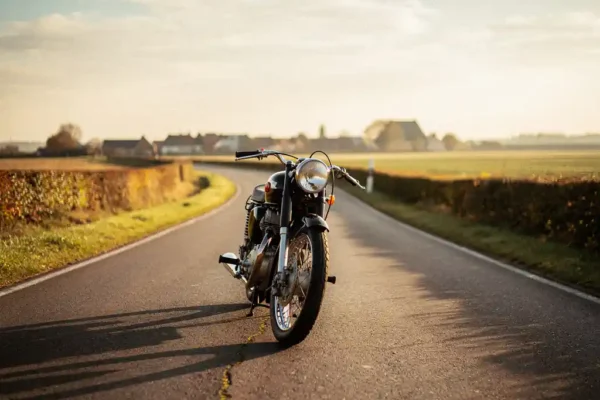 Klassieke motorfiets op verlaten Belgische landweg in gouden middaglicht, kalender met drie maanden op de voorgrond, lange schaduwen op het asfalt.