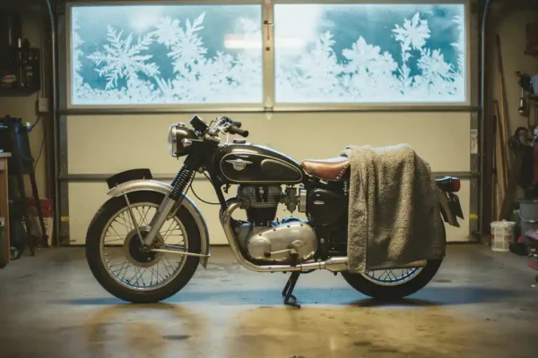 Klassieke motorfiets met stoflaag op de tank in garage, winterhoes half van het zadel, bevroren raam op achtergrond.
