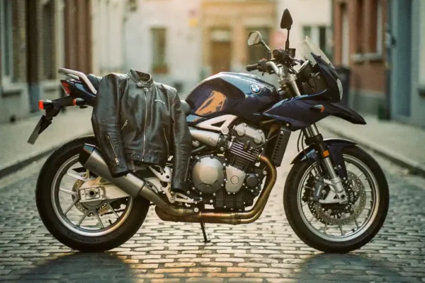 Glanzende marineblauwe motorfiets op een Belgische kasseienstraat bij gouden uurlicht, met een leren rijjas over het zadel.