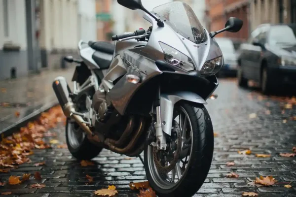 Beschadigde motorfiets op natte Belgische kinderkopjes met gebarsten spiegel en gekraste kuip, omgeven door herfstbladeren.