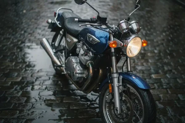 Regendruppels op een motorfiets geparkeerd op natte Belgische kasseien, met chroom details en een bewolkte lucht op de achtergrond.