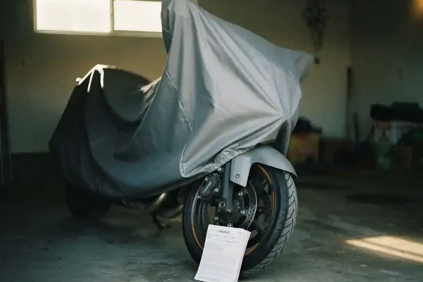 Afgedekte motorfiets in schemerige garage met verzekeringsdocument tegen het voorwiel, zacht daglicht door smal raam.