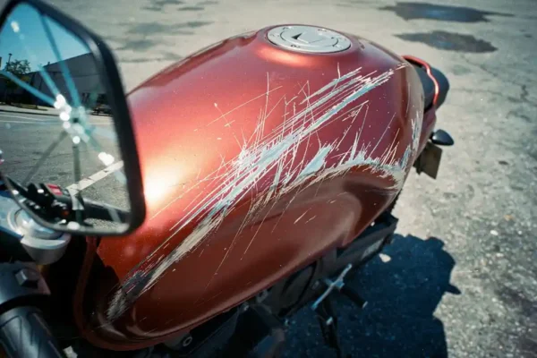 Beschadigde motorfiets met krassen en sleutelsporen op metallic rode tank, geparkeerd op leeg stedelijk parkeerterrein.