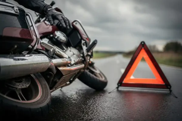 Motorrijder met gehandschoende hand op gevallen toermotor langs natte Belgische weg, gevarendriehoek op asfalt.