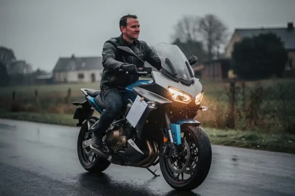 Motorrijder op natte Belgische weg met verzekeringsDocument dat uit jaszak glijdt, bewolkte lucht op de achtergrond.