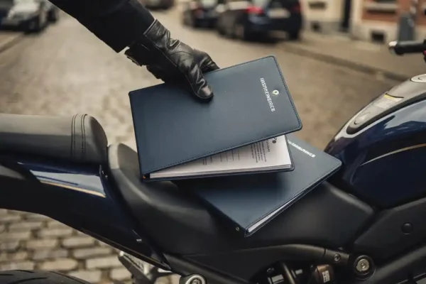 Motorrijder in handschoen legt nieuwe verzekeringsdocumenten op motorzadel in Belgische straat, symboliseert overstap van motorverzekering.