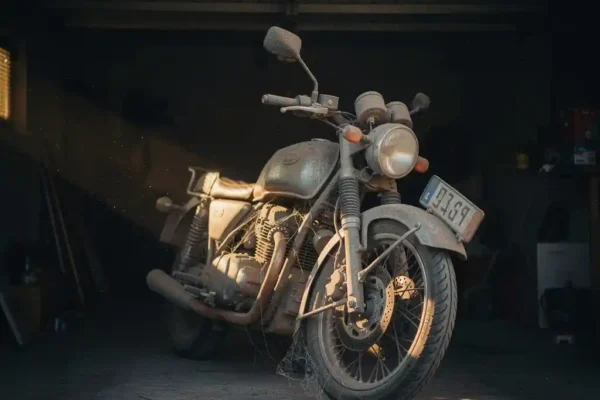 Stoffige motorfiets met Belgisch kenteken in een schemerige garage, met spinnenwebben in de spaken en een dikke laag stof op het stuur.